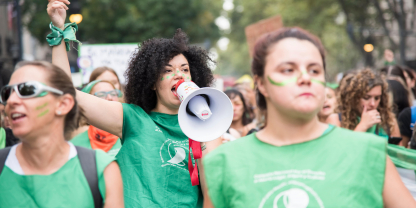 A young black woman speaking into a megaphone with a fist in the air. She is wearing a green top used as the symbol of the pro-choice movement in Latin America.  