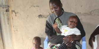 A Kenyan woman sitting down, holding a smiling baby. Another one of her young children is standing next to her.