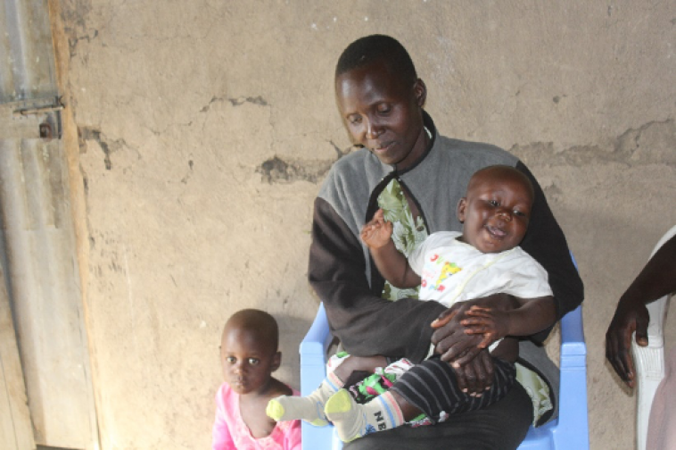 A Kenyan woman sitting down, holding a smiling baby. Another one of her young children is standing next to her.